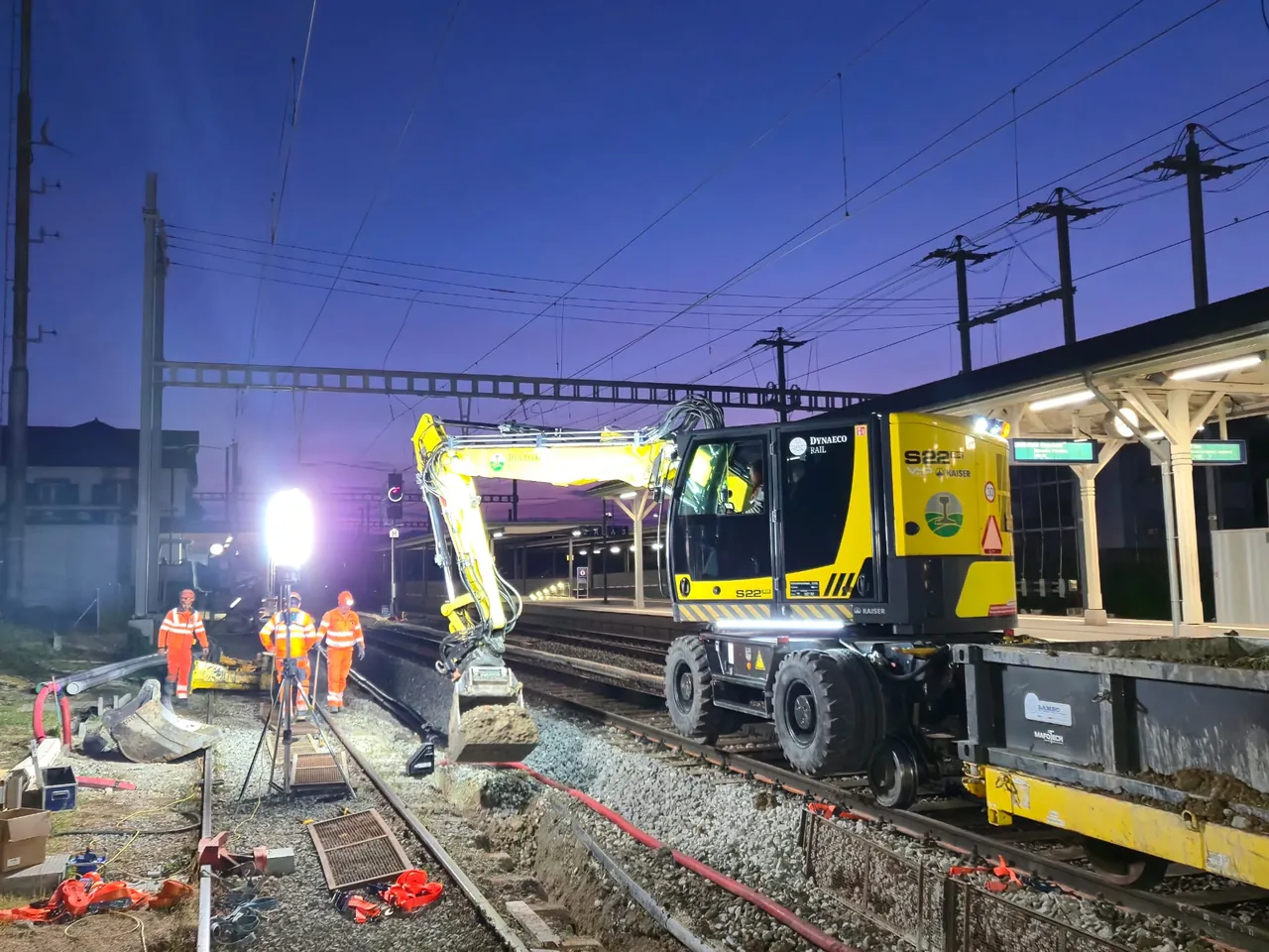 Chantier ferroviaire de nuit près d'une gare, avec engin et balisage
